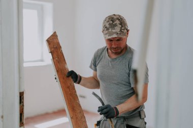 A young caucasian man in a uniform and gray textile gloves with real emotions on his face holds a crowbar and a torn board from a doorway in his hands, close-up side view with selective focus. Construction work concept.