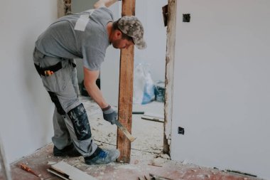 A young Caucasian man in a uniform and gray textile gloves standing sideways and in an inclination knocks out a nail from a dismantled board with a crowbar and copy space on the right, close-up side view with selective focus. Construction work concep