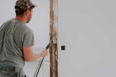 Üniformalı ve gri tekstil eldivenli genç beyaz bir adam elinde levyeyle arkadan dönük duruyor kapının önündeki tahtadaki eski alçıyı söküyor ve sağ tarafta seçici bir odak noktası var. İnşaat iki