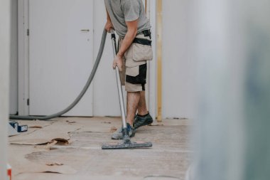 A young caucasian brunette man unrecognizable in work clothes on the left is vacuuming the floor with a construction vacuum cleaner in a room in an old house where renovations are underway, close-up view from the bottom with selective focus.Construct