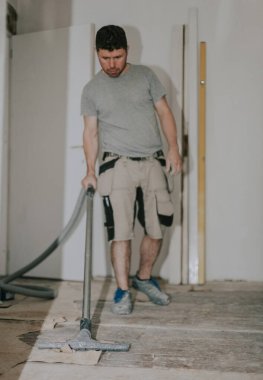 Young caucasian brunette man in work clothes standing in front vacuums the floor with a construction vacuum cleaner in a room in an old house where renovations are taking place, bottom view close-up with selective focus.Construction work concept.