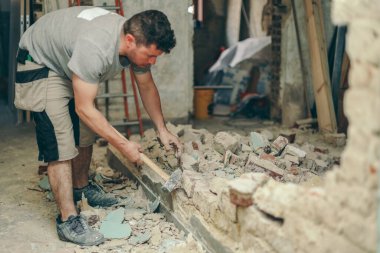 Young caucasian brunette man with a beard in work clothes breaks a brick wall with a sledgehammer in an old house,side view close-up with selective focus.Construction work concept.