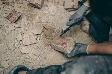 A young caucasian man in black work clothes sits on a small chair on the right and cleans the bricks from dirt and old cement with an ax, top view close-up.Construction work concept.