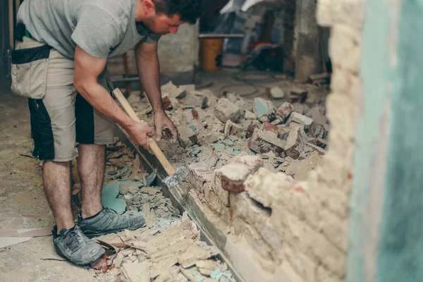 Young caucasian brunette man with a beard in work clothes breaks a brick wall with a sledgehammer in an old house,side view close-up with selective focus.Construction work concept.