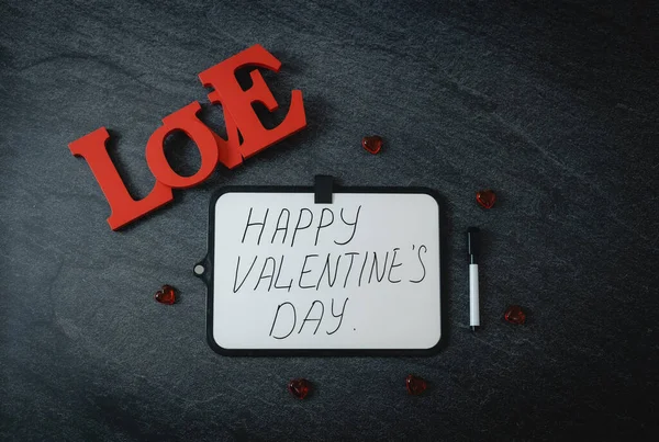 One marker board with handwritten lettering: Happy valentine\'s day,wooden word love and red glass hearts on black stone background,flat lay closeup. Valentine\'s day concept.