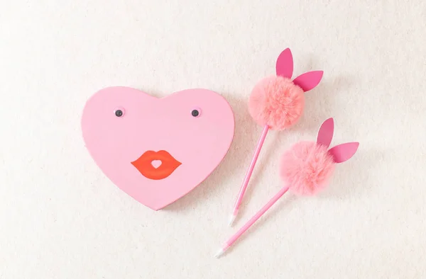 One heart gift box with eyes, paper mouth, smile and two pens with a fluffy head and bunny ears lie in the center on a natural beige stone background, flat lay close-up. Valentine\'s day concept, happy easter.