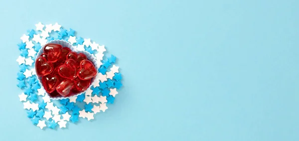 One muffin tin with glass red hearts and candy sprinkles stars around lie on the left on a blue background with copy space on the right, flat lay close-up. Valentine's day concept.