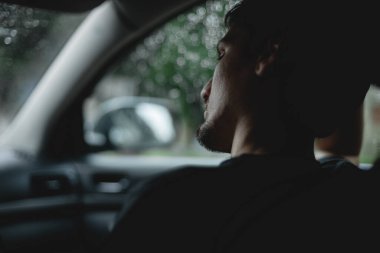 Portrait of one young handsome caucasian brunette guy with a beard sits thoughtfully in a car in the rain, side view close-up with depth of field.