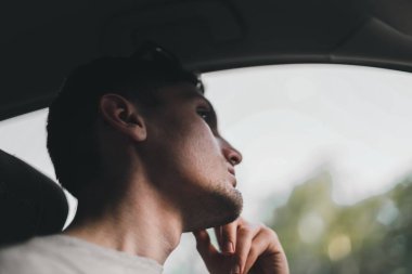 Portrait of a young handsome caucasian brunette guy with a beard and sunglasses thoughtfully sitting behind the wheel of a car in a traffic jam and holding his chin with his hand, close-up view from below.
