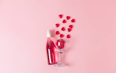One bottle of wine and a glass with glass hearts pouring out of it lies in the center on a pink background with copy space on the sides, flat close-up. The concept of love and the holiday of Valentine's Day.