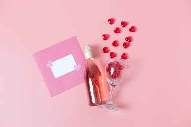 One envelope, a bottle of wine and a glass with glass hearts pouring out of it lies in the center on a pink background, flat close-up. The concept of love and the holiday of Valentine's Day.