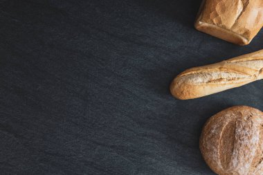 Two loaves and one french baguette lie to the right on a black stone background with copy space on the left, flat lay close-up. Bread baking concept.