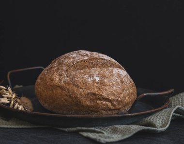 One round rye bread in a vintage metal plate with a kitchen napkin and ears lie on a black stone background, close-up side view.Bread baking concept.