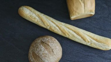 Two breads of different sotres and forts with one French baguette lie in the center on a black stone background with tracking to the right, flat lay close-up. Bread baking concept.