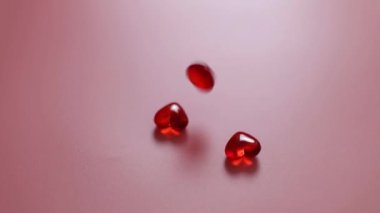 Falling one by one red small glass hearts and bouncing on a pink background, close-up side view. Concept of love, valentine's day.