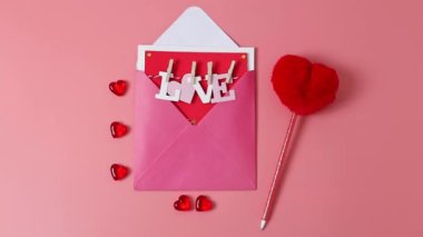 One envelope with a letter inside with a paper word love on a rope with clothespins, a red decorative glass heart and a pen with a fluffy heart lie on a pink background with a zoom in closeup. The concept of love, valentine's day.