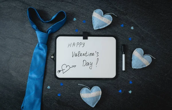 One empty marker board with the inscription: happy valentine\'s day, blue tie, three textile hearts and shiny confetti on a black stone background, flat lay close-up. Valentine\'s day concept.