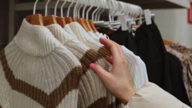 The hand of a young caucasian girl sorts through sweaters on a coat hanger hanging on a stand in a store, close-up side view. Offline shopping concept.