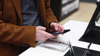 Young caucasian unrecognizable man in a brown coat inspects a new mobile phone on the counter of a store,side view close-up in slow motion,side view close-up.Smartphone shopping concept.