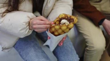 A young caucasian girl in a white puffy jacket and jeans sits on a white bench in the mall and eats a delicious waffle pimply dessert with filling, close-up side view.Concept of eating dessert.