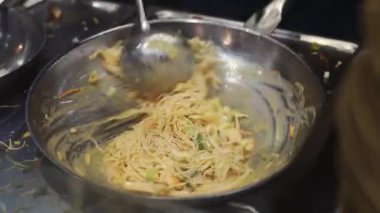The hand of a young caucasian unrecognizable girl stirs pasta with vegetables in a frying pan with a ladle while cooking wok food in a mall diner, close-up side view.Concept of cooking wok food.