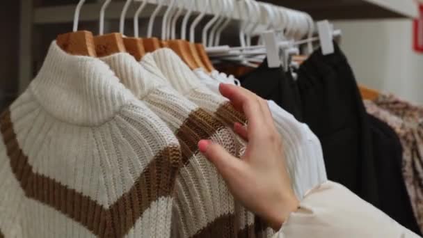 一个年轻的高加索女孩的手通过挂在商店的架子上的外套衣架上的毛衣进行分类 这是一个近距离的侧面视图 离线购物概念 — 图库视频影像