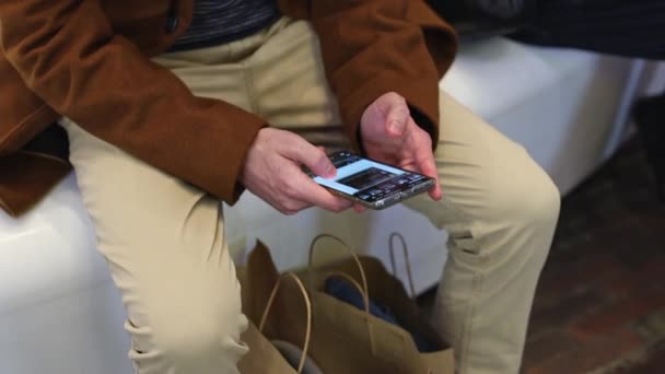 身穿棕色外套的年轻而难以辨认的高加索人坐在购物中心的白色长椅上 一边用手机等着他的妻子 一边从放大的视野中看到近景 用科技概念 在购物中心等着 — 图库视频影像