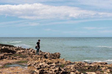A handsome caucasian young brunette guy in jeans and a t-shirt stands on a rocky shore looking at the north sea in Etretat in France, close-up view from above. People and nature concept.