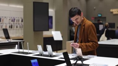 Young caucasian recognizable man in a brown coat examines a smartag for a mobile phone, standing behind the counter of a store, close-up from the side in slow motion, close-up from the side. Smartphone shopping concept.
