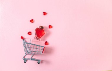 One small cart, a bottle in the shape of a heart with red perfume and a glass decoration of hearts lie on the left on a pink background with copy space on the right, flat lay close-up. Valentine's day concept, holiday shopping.