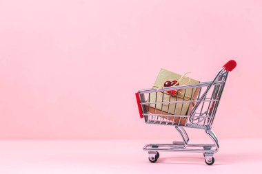 One small trolley and craft gift box rewound with jute thread with red glass heart lie on the right on a pink background with copy space on the left, close-up side view. Valentine's day concept, holiday shopping, holiday gifts.