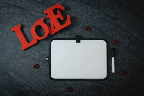 One empty marker board, wooden word love and red glass hearts on black stone background with copy space, flat lay closeup. Valentine\'s day concept.