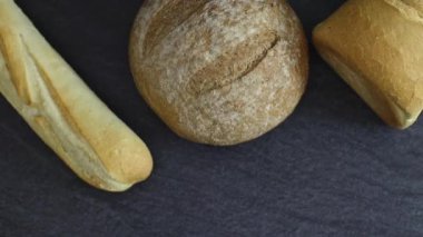 Two breads of different varieties with one French baguette lie against a background of black stone with tracking to the right, close-up view from above. The concept of baking bread.