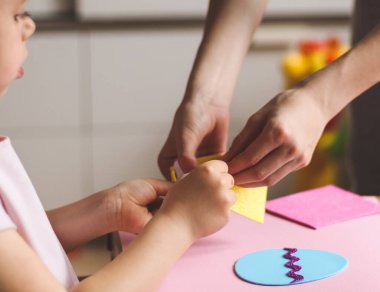 The hands of a little caucasian girl hold a yellow sticker, and mom's hands help peel off a strip for felt eggs while sitting at a children's table with a craft object set on a pink background, flat lay close-up.