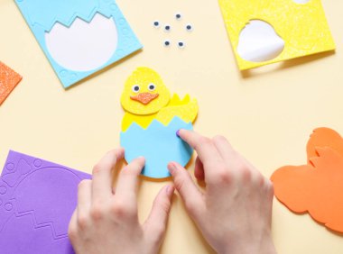 Hands of a caucasian teenage girl sticking a sticker of a blue shell with a zigzag tear on a yellow chicken felt with her fingers, sitting at a children's table with a set of handmade items on a pale yellow background with selective focus, flat lay c