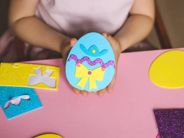 The hands of a little caucasian girl show the end result of a homemade felt easter egg, sitting at a children\'s table with a set of crafts on a pink background with depth of field, close-up side view. The concept of crafts, diy, needlework, diy, chil