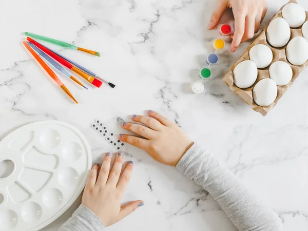 Children\'s hands are stretched out to glued eyes on a marble table with white eggs in a cardboard box, brushes, acrylic paint and a palette for preparing their own hands for the Easter holiday, flat lay close-up. Concept diy, artisanal, at home, chil