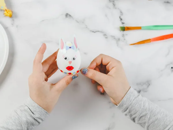 Hands of a caucasian girl in a gray turtleneck holding an easter bunny egg and sticking a colored sticker with her finger while sitting at a marble table for diy preparation for the easter holiday, flat lay close-up. The concept of crafts, needlework