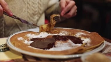 Young caucasian unrecognizable man eating sweet pancake dessert with chocolate with a knife and fork while sitting at a table in a restaurant, close-up side view. Men's lifestyle concept.