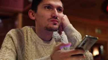 A young caucasian handsome brunette man sits at a table in a restaurant with a mobile phone in his hands, looking attentively to the side and emotionally talking, close-up side view. Men's lifestyle concept, using technology.