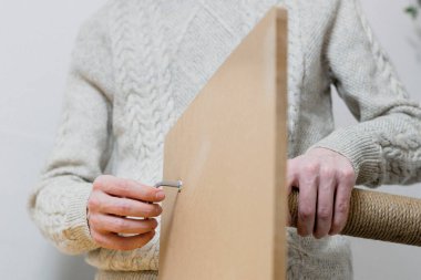 Young caucasian man in a knitted sweater and jeans manually drives a screw into a board while assembling a cat scratching post, standing at a table against a white wall, close-up view from below. Concept of assembling a scratching post, cat furniture