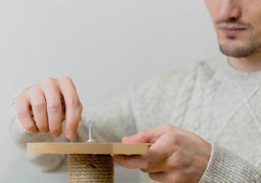 Young handsome caucasian man in a knitted sweater manually tightens a screw into a board while assembling a cat scratching post against a white wall, close-up side view. Concept of assembling a scratching post, cat furniture.