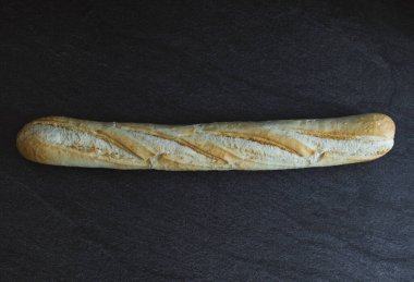 One french white wheat baguette lie in the center on a black stone background with copy space on the sides,flat lay closeup.Bread baking concept.