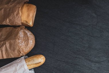 Two breads and one french baguette in paper bags lie on the left on a black stone background with copy space on the right, flat lay close-up. Bread baking concept.
