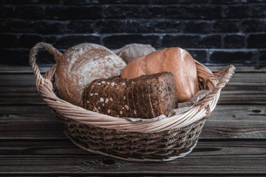 Three different varieties and shapes in an oval wicker basket with handles lie in the center on a brown wooden table against a blurred black brick wall, close-up side view. The concept of baking bread.