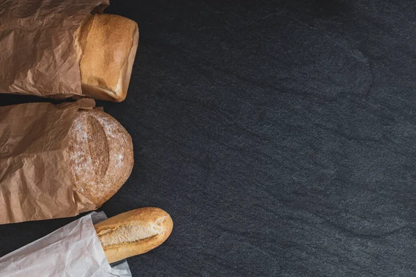 Two breads and one french baguette in paper bags lie on the left on a black stone background with copy space on the right, flat lay close-up. Bread baking concept.