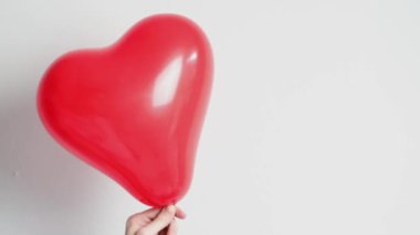 Hand of young caucasian man waving left and right red heart balloon on white wall background,side view close-up in slow motion.Valentine's day concept.