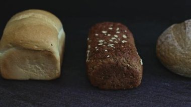 Three loaves of different varieties, sizes and shapes lie on a black stone background, close-up side view with tracking to the right. The concept of baking bread.