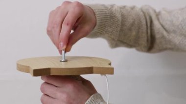 Hands of a young caucasian unrecognizable man in a knitted sweater hand-twisting a screw into a creative wooden cat scratching post, standing at a table in a room, close-up side view.Concept of assembling a scratching post, furniture.