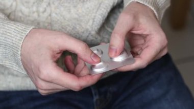 The hands of a young caucasian man in jeans and a knitted sweater sitting on a chair squeeze a white pill out of a gray package, close-up side view. Concept of medicine and healthcare.